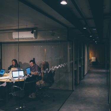 Office corridor at night with people in room behind glass wall